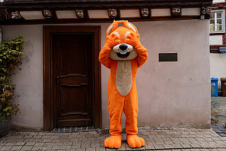 Das Maskottchen der hessischen Turnjugend steht vor dem Haus der sieben Laster in Limburg und hält sich die Augen zu.
