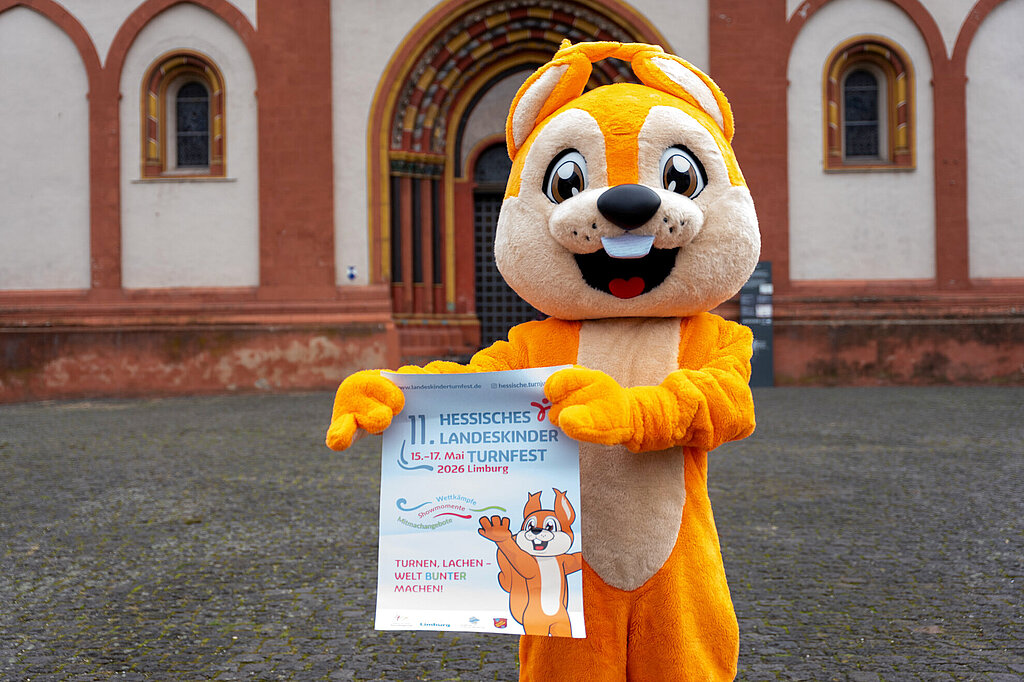 Das HTJ-Maskottchen Freddy hält ein Plakat vom Landeskinderturnfest in den Händen.