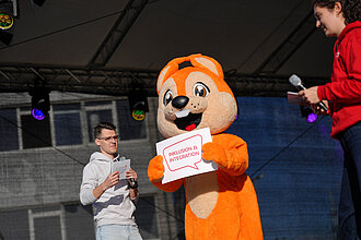 Das Maskottchen Freddy, ein Eichhörnchen, hält auf der Bühne ein Schild hoch.
