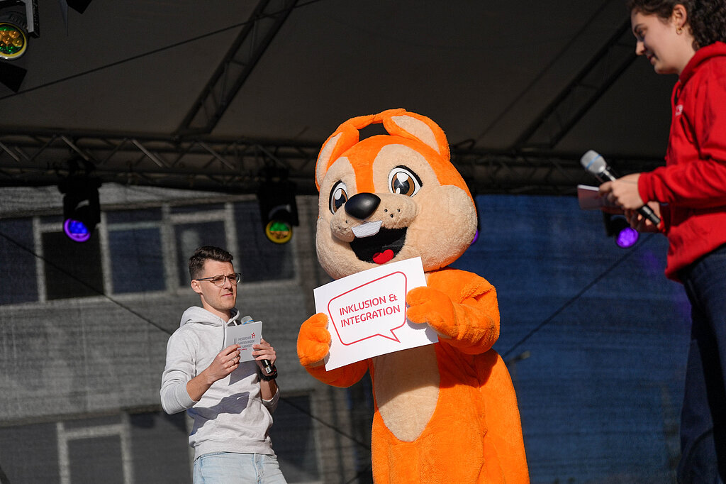 Das Maskottchen Freddy, ein Eichhörnchen, hält auf der Bühne ein Schild hoch.