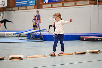 Kleines Mädchen balanciert über verschiedene kleine Geräte in einer Sporthalle.