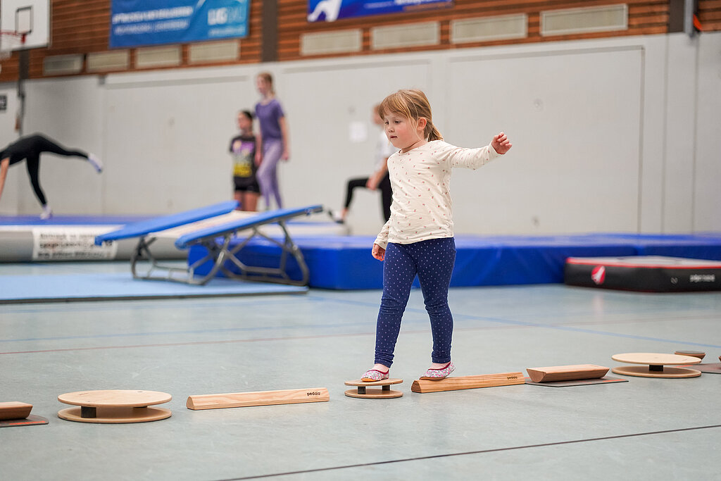 Kleines Mädchen balanciert über verschiedene kleine Geräte in einer Sporthalle.