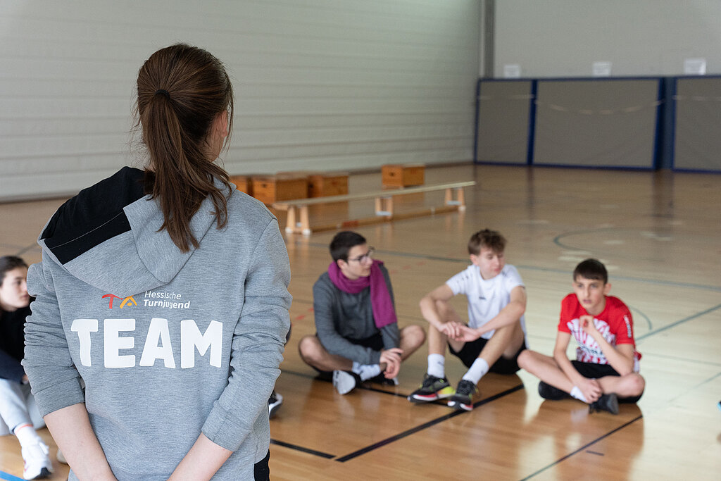 Eine Junge Frau mit Jacke erklärt Schülern etwas in einer Sporthalle.