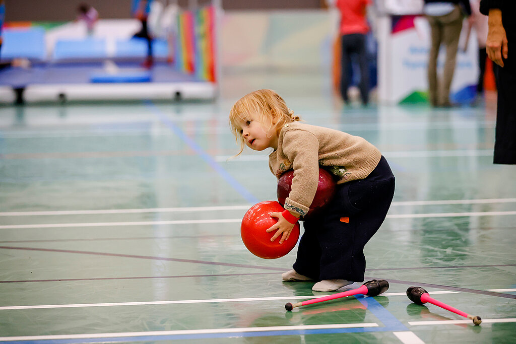 Ein kleines Kind hebt in einer Sporthalle einen roten Ball auf.